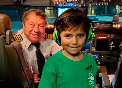 A boy wearing green headphones and a green t-shirt standing in the cockpit of a plane. The pilot sits behind him.