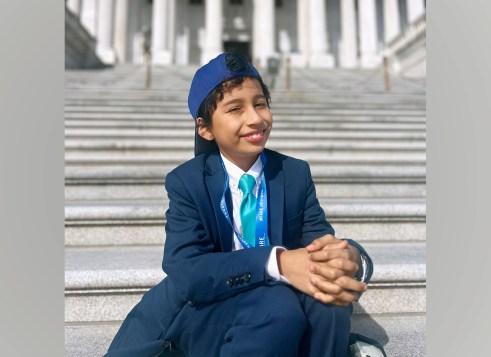 Kid in suit and blue cap sits smiling on Capitol steps.