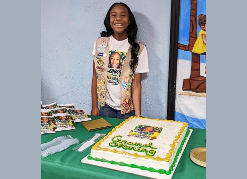 A girl wearing a Girl Scouts vest stands smiling behind a large “Grand Opening” cake displayed on a table at an event.