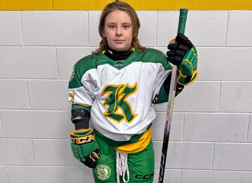 A young hockey player in a green and white uniform stands against a wall, holding a hockey stick and looking at the camera.