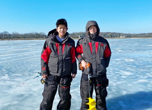 Two kids wearing winter gear stand on a frozen lake. One holds a fishing rod, the other drill with an ice auger.