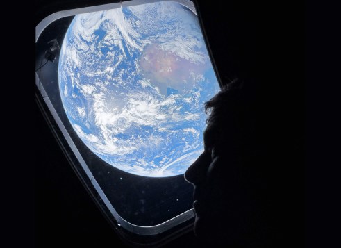 A person looks out a spacecraft window at Earth, which appears bright and blue against the dark background of space.