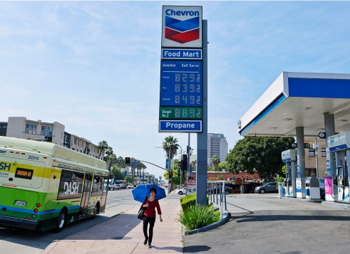 A gas station sign shows fuel prices as a bus and a person with an umbrella pass by on the street.