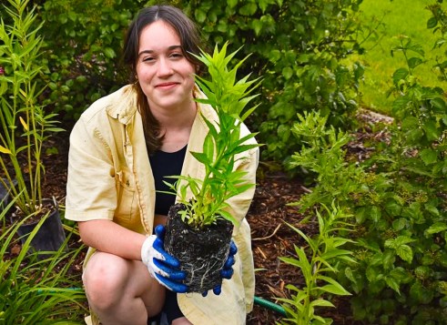 A girl kneels in a garden, smiling and holding a young plant with roots.