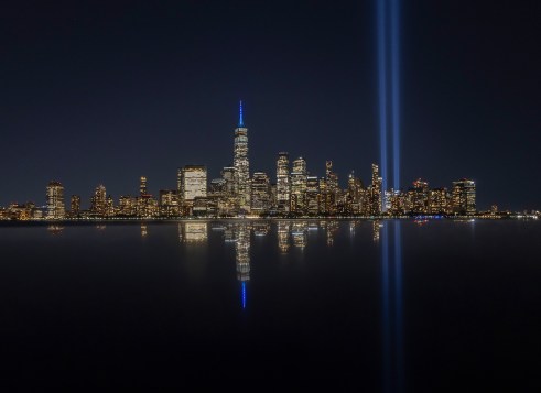 A city skyline glows at night as two beams of blue light shine straight into the sky.