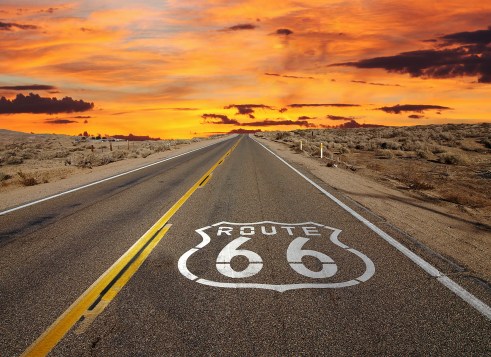 A highway marked with a large route shield stretches toward the horizon beneath a fiery sunset.